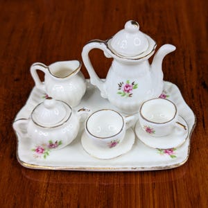 May include: A miniature porcelain tea set on a rectangular tray. The set includes a teapot, creamer, sugar bowl, and two teacups with saucers. Each piece is white with gold trim and pink rose accents. The tray has a scalloped edge.