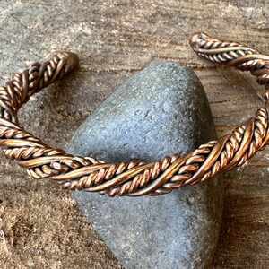 May include: A copper cuff bracelet with a braided rope design. The bracelet is a warm, reddish-brown color and appears to be handmade. The cuff is resting on a gray stone against a wooden background.