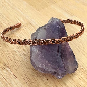 May include: A copper wire bracelet with a twisted design. The bracelet is on a purple amethyst crystal.