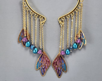 Könnte beinhalten: Goldfarbene Ohrklemmen mit herabhängenden Ketten, verziert mit lila und blauen flügelförmigen Anhängern und türkisfarbenen Perlen. Die Ohrklemmen haben ein geschwungenes Design.