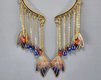 Könnte beinhalten: Goldener Ohrring mit herabhängenden Ketten und blauen und orangen Perlen. Die Ketten sind mit kleinen Schmetterlingsanhängern verziert.