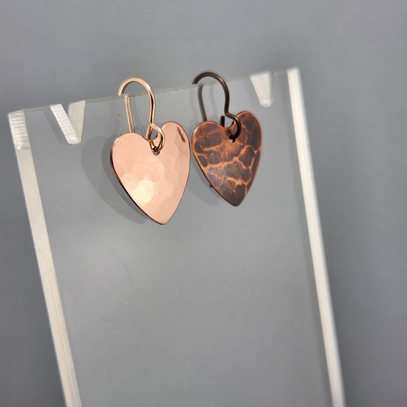 May include: Two heart-shaped earrings. One is a hammered copper color, the other is a darker, textured brown. Both have simple ear wires. The earrings are displayed on a clear stand against a gray background.