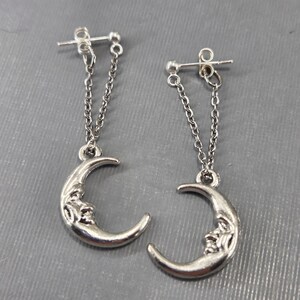 May include: A pair of silver crescent moon earrings with a face on the moon. The earrings have a silver chain that hangs from the moon.