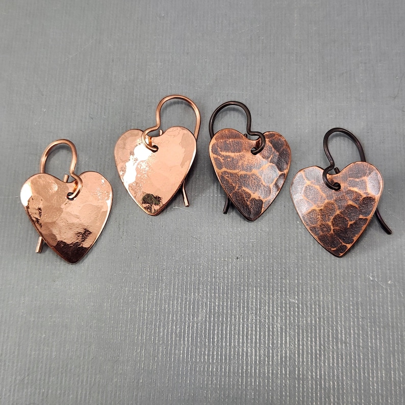 May include: Four heart-shaped earrings. Two are copper-colored with a hammered texture, and two are a darker, bronzed color with a textured pattern. Each earring has a curved hook for pierced ears. The earrings are displayed on a gray surface.