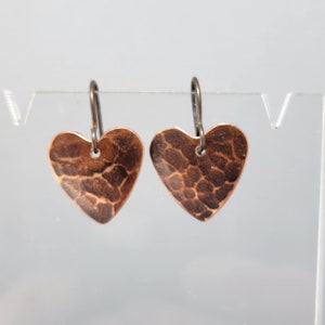May include: A pair of copper heart-shaped earrings. The earrings have a hammered texture and are suspended from dark metal ear wires. The hearts are a warm, reddish-brown color, and the overall design is simple and elegant.