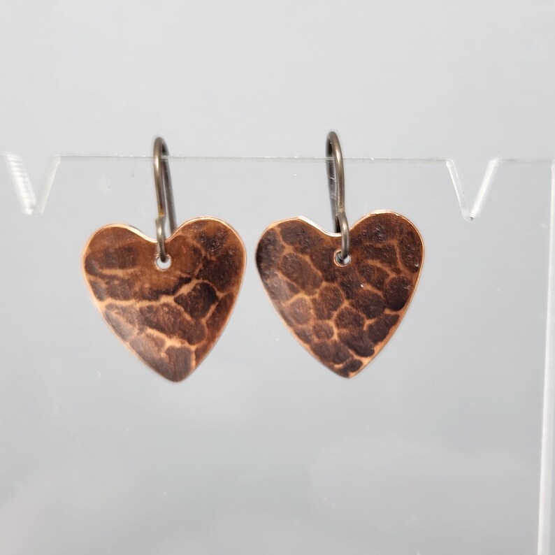 May include: A pair of copper heart-shaped earrings. The earrings have a hammered texture and are suspended from dark metal ear wires. The hearts are a warm, reddish-brown color, and the overall design is simple and elegant.