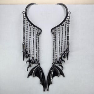 May include: A pair of black ear cuffs featuring curved frames with multiple dangling chains. Each cuff is adorned with small black beads, pointed spikes, and a prominent bat wing charm at the base, creating a layered, symmetrical design.