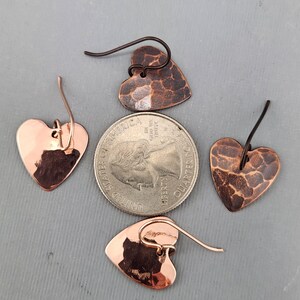 May include: Four copper heart-shaped earrings. Two earrings have a hammered texture, and two are smooth. The earrings are displayed with a quarter for size reference. The ear wires are copper and dark brown.