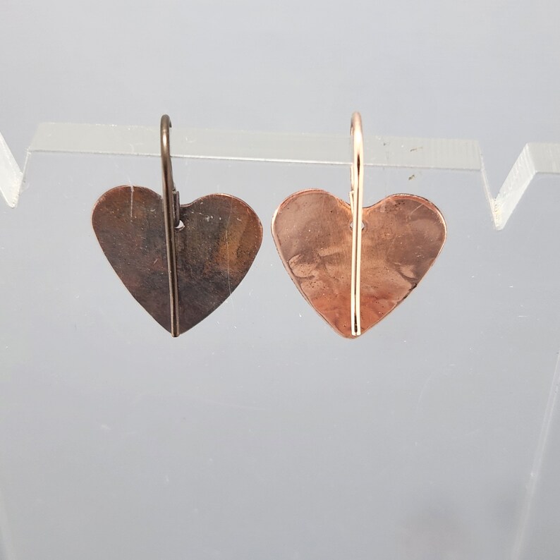 May include: A pair of heart-shaped earrings. One earring has a dark brown finish, while the other has a copper finish. Both earrings have a wire hook for pierced ears. The earrings are displayed against a light gray background.
