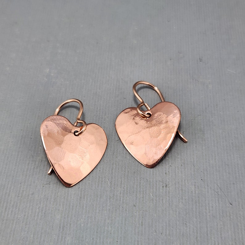 May include: A pair of copper heart-shaped earrings with hammered texture. The earrings have a simple hook design and a warm, reddish-brown color. The earrings are displayed on a gray surface.