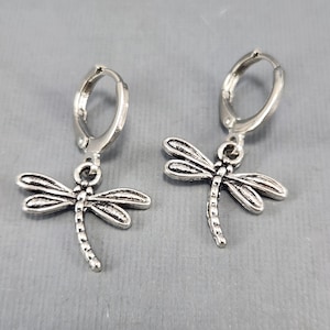May include: A pair of silver hoop earrings with dragonfly charms. The dragonflies have detailed wings and a long, slender body.