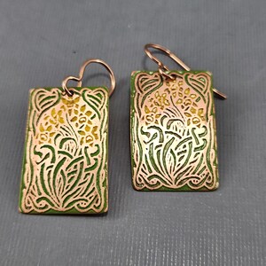 May include: A pair of green and gold earrings with a floral design. The earrings are made of metal and have a rectangular shape.