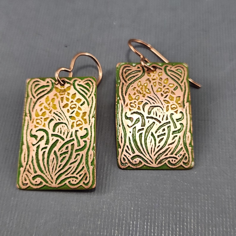 May include: A pair of green and gold earrings with a floral design. The earrings are made of metal and have a rectangular shape.
