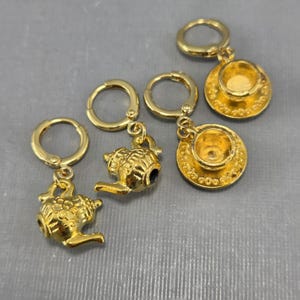 May include: Gold-coloured earrings with teapot and teacup charms. The earrings have a hoop closure and feature detailed miniature teapot and teacup pendants. The teacups have a saucer base. The earrings are displayed on a grey surface.
