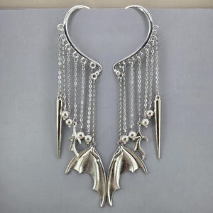 May include: A pair of silver-toned ear cuffs featuring a curved upper frame with multiple dangling chains. Each cuff includes small metallic spheres, a pointed spike charm, and a bat wing charm suspended from the chains, creating a layered design.