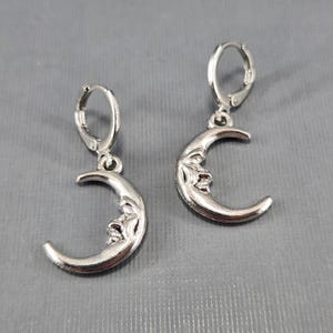 Pode incluir: Um par de brincos de argola de prata com um pingente de lua crescente. O pingente de lua tem um rosto.