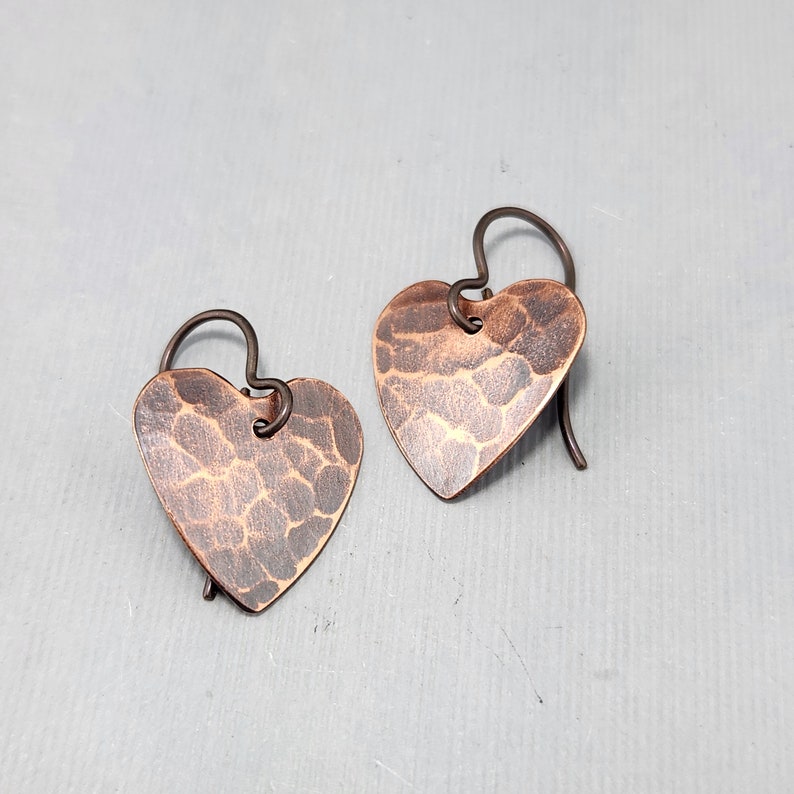 May include: A pair of handcrafted copper heart earrings. Each earring features a hammered texture and a dark, antiqued finish. The heart-shaped pendants are suspended from simple ear wires, creating a rustic and romantic aesthetic.