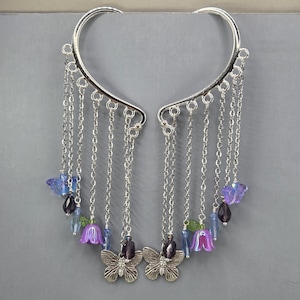 May include: A pair of silver-toned ear cuffs featuring multiple dangling chains. The chains are adorned with small purple and blue glass butterfly beads, bell-shaped flower beads, and metallic butterfly charms at the bottom of the strands.