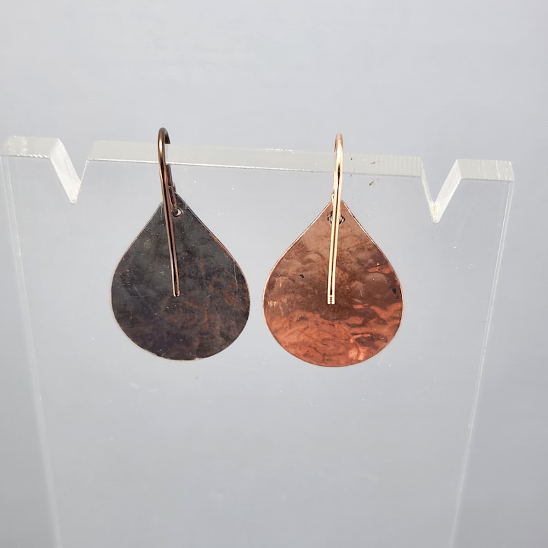 May include: A pair of teardrop-shaped earrings with a hammered copper finish. One earring is a darker copper color, while the other is a lighter copper color.