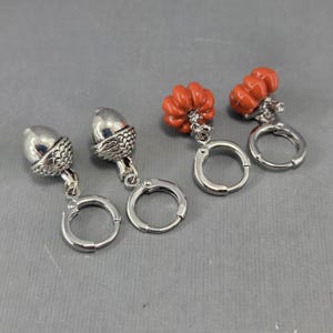 Può includere: Orecchini a forma di ghianda e zucca arancione color argento. Gli orecchini a ghianda hanno un cappuccio dettagliato e pendono da piccole chiusure a cerchio. Gli orecchini a zucca sono di un arancione vibrante e pendono anche da chiusure a cerchio argentate.