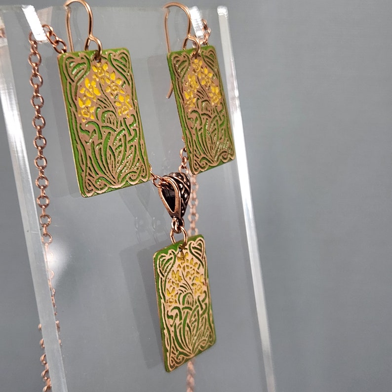 May include: A green and gold necklace and earrings set with a floral design. The pendant and earrings are rectangular and have a delicate floral pattern with yellow accents.