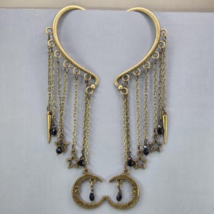 May include: A pair of antique brass-toned ear cuffs featuring a curved frame with multiple dangling chains. The chains are adorned with small open-work star charms, dark faceted beads, and a crescent moon charm at the base of each piece.