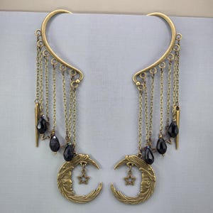 May include: A pair of antique gold-colored ear cuffs. Each cuff features a crescent moon with a face, a dangling star, black beads, and chains. The cuffs have a curved design that fits over the ear.