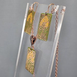 May include: A copper necklace and earrings set with green and yellow enamel floral designs. The necklace has a pendant with a rectangular shape and the earrings are also rectangular.