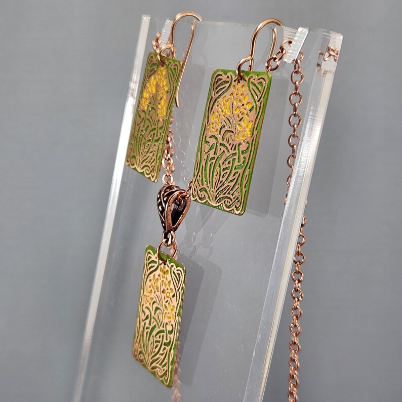 May include: A copper necklace and earrings set with green and yellow enamel floral designs. The necklace has a pendant with a rectangular shape and the earrings are also rectangular.