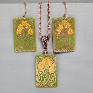 Puede incluir: Un conjunto de collar y pendientes de esmalte verde y amarillo con un diseño floral. El colgante y los pendientes son rectangulares y presentan un diseño floral estilizado con flores amarillas y hojas verdes.