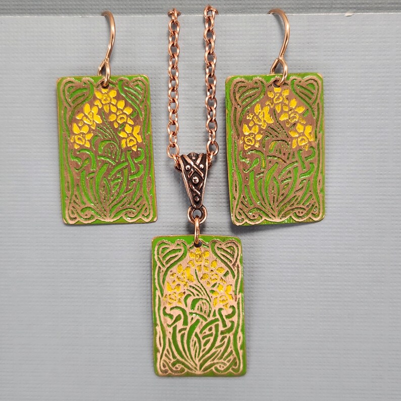 May include: A green and yellow enamel necklace and earrings set with a floral design. The pendant and earrings are rectangular and feature a stylized floral design with yellow flowers and green leaves.