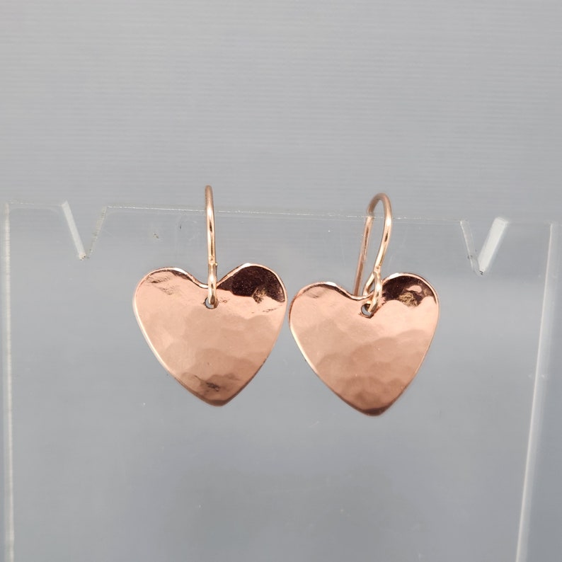 May include: A pair of rose gold heart-shaped earrings with a hammered texture. The earrings hang from delicate ear wires, showcasing a simple yet elegant design. The hearts are symmetrical and have a slightly reflective surface.