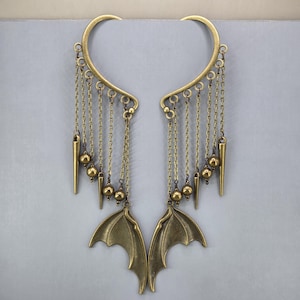May include: A pair of antique brass ear cuffs featuring a curved upper frame with multiple dangling chains. Each chain ends in either a small metal bead or a pointed spike. At the base of each cuff hangs a detailed bat wing charm in brass.