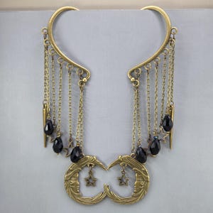 May include: A pair of antique brass ear cuffs featuring a curved frame with multiple dangling chains. Each chain is adorned with black teardrop beads and small star charms. The bottom of each cuff showcases a crescent moon with a face design.