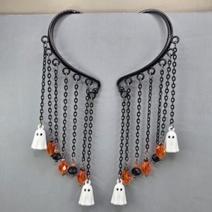 May include: A pair of black metal ear cuffs featuring a curved frame with multiple dangling black chains. Each chain is accented with small orange and black beads, and the ends are finished with small white ghost charms with black eyes.