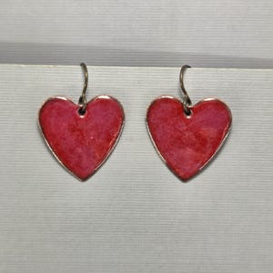 Op de afbeelding: Een paar hartvormige oorbellen. Elke oorbel heeft een felrood hart met een metallic zilveren rand en een eenvoudige haak voor het oor. De oorbellen worden gepresenteerd tegen een neutrale achtergrond.