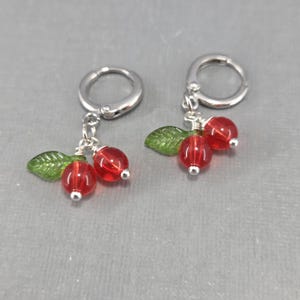 May include: A pair of silver hoop earrings with dangling red cherry charms. Each charm features two red glass beads, a green leaf, and a silver bead. The earrings are displayed on a gray surface.