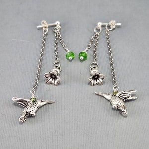 May include: Silver dangle earrings with two chains, a green crystal bead, a silver flower charm, and a hummingbird charm.