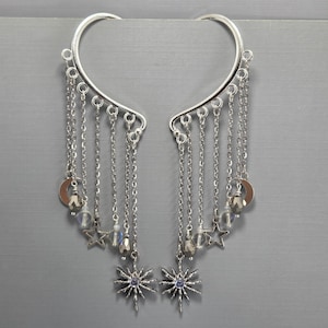 May include: Silver ear cuffs with dangling chains, stars, beads, and sunburst charms. The ear cuffs curve around the ear and feature multiple chains with decorative elements. The sunburst charms have a small purple stone in the center.
