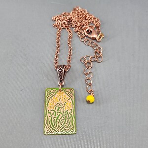 May include: A copper necklace with a green and yellow enamel pendant featuring a floral design. The pendant is rectangular and has a detailed border. The necklace has a lobster clasp and a small yellow bead.