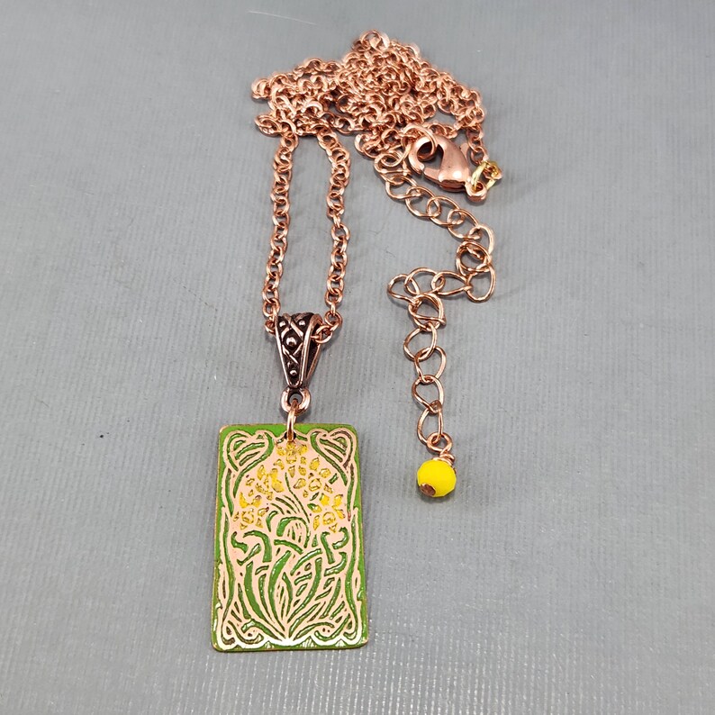 May include: A copper necklace with a green and yellow enamel pendant featuring a floral design. The pendant is rectangular and has a detailed border. The necklace has a lobster clasp and a small yellow bead.