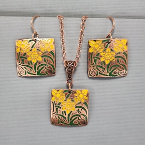 Puede incluir: Un conjunto de collar y pendientes de cobre con un colgante cuadrado y pendientes con un diseño floral con narcisos amarillos y hojas verdes.