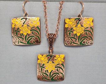 May include: A copper necklace and earrings set with a square pendant and earrings featuring a floral design with yellow daffodils and green leaves.