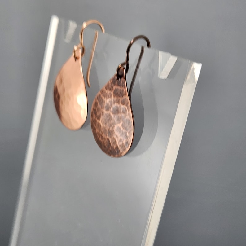 May include: A pair of hammered copper teardrop earrings with a shiny finish. The earrings are hanging from a clear acrylic display stand.