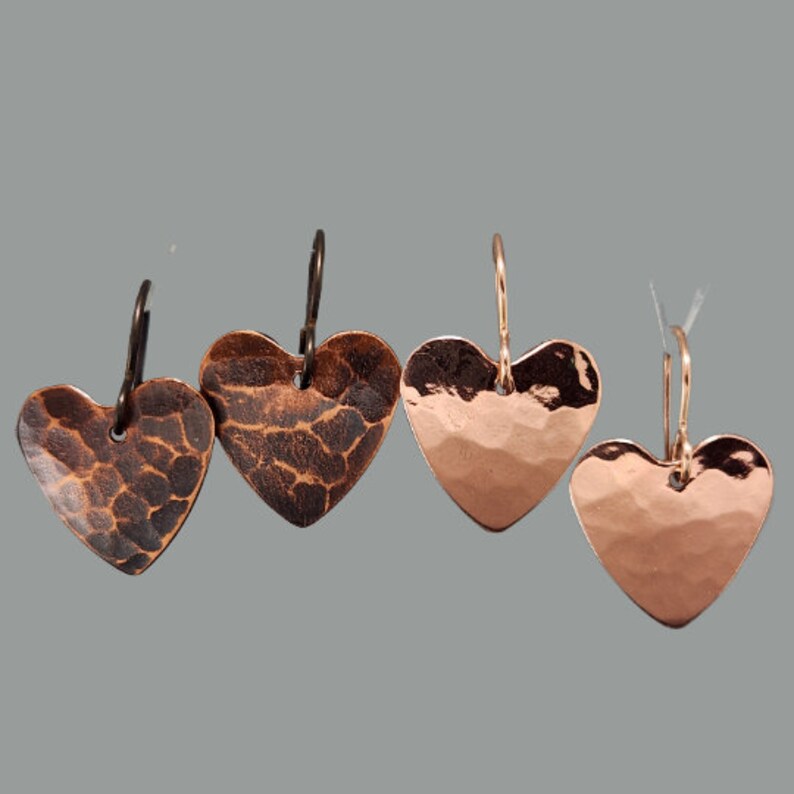 May include: Four pairs of copper heart-shaped earrings. Two pairs have a textured, dark brown finish, while the other two have a smooth, polished copper appearance. The earrings hang from ear wires.