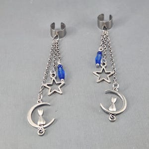 May include: A pair of silver ear cuffs with dangling charms. Each cuff has three chains with a blue bead, a star charm, and a crescent moon charm with a cat silhouette.