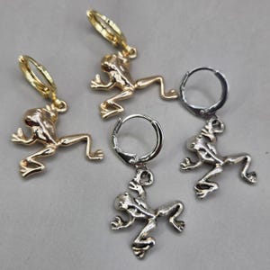 Pode incluir: Quatro brincos em forma de sapo. Dois são dourados e dois prateados. Cada sapo está numa pose de salto e pende de um pequeno brinco de argola. Os brincos são exibidos numa superfície cinzenta.