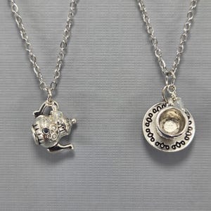 May include: Two silver-toned necklaces. One features a teapot charm, the other a teacup and saucer charm. Both charms are detailed and suspended from delicate silver chains. The background is a solid gray.