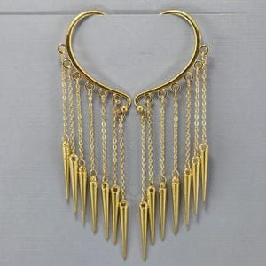 May include: A pair of gold-colored ear cuffs. Each cuff has a curved top with dangling chains and cone-shaped pendants. The ear cuffs are displayed against a gray background.
