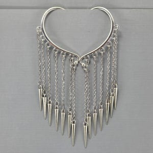 May include: Silver-toned ear cuff with a curved top and dangling chains. Each chain ends with a cone-shaped spike. The ear cuff is designed to be worn on the ear and is displayed against a gray background.
